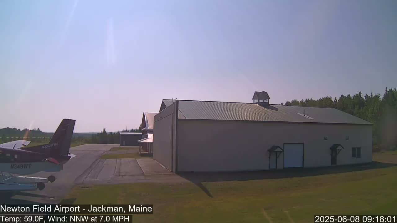 A small airplane sits on a tarmac next to a large hangar on a sunny day with a light north-northwest wind.