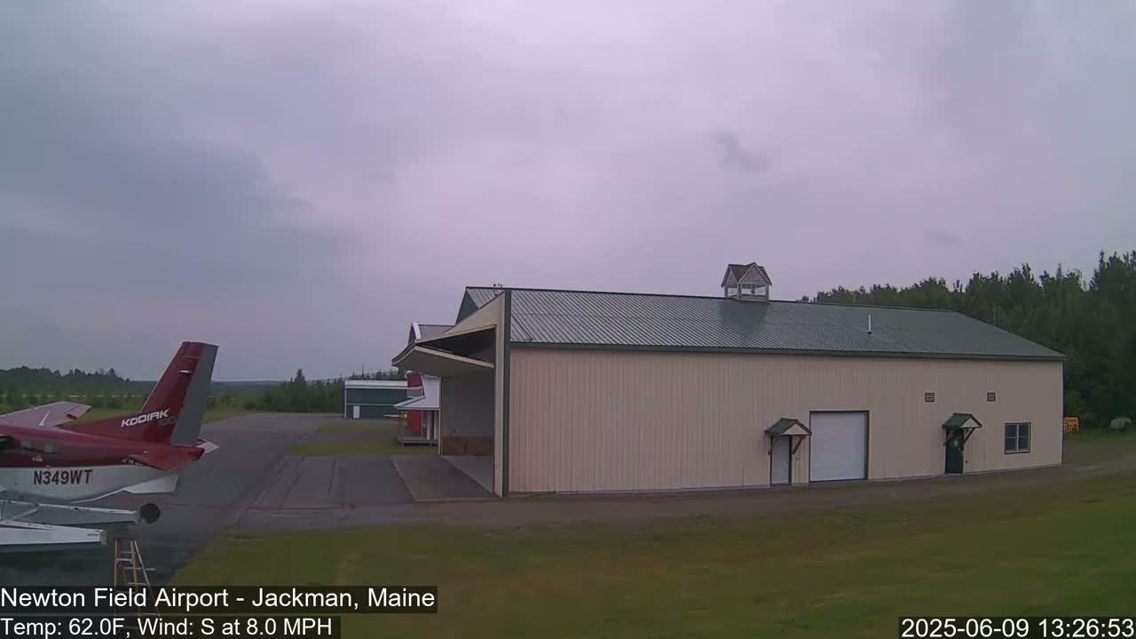 A red airplane is parked on an airport tarmac next to a large beige hangar under an overcast sky.