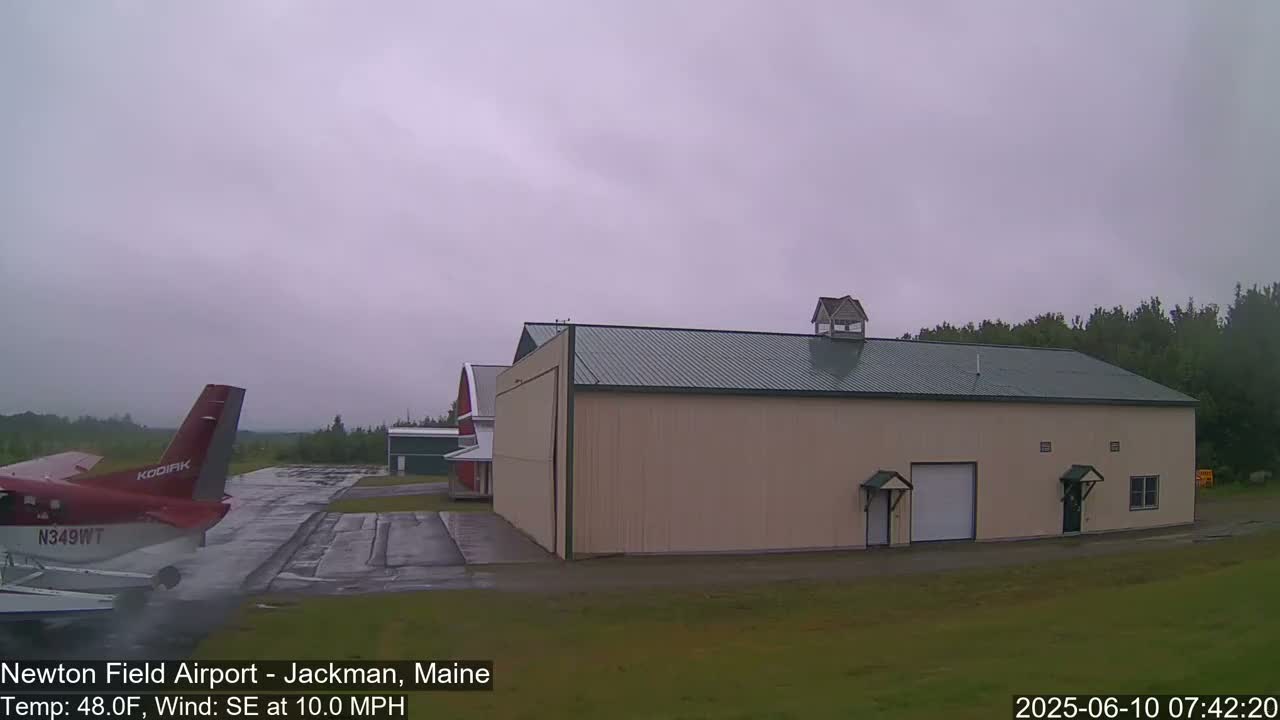 A small airplane sits on a tarmac next to a large tan hangar under an overcast sky.