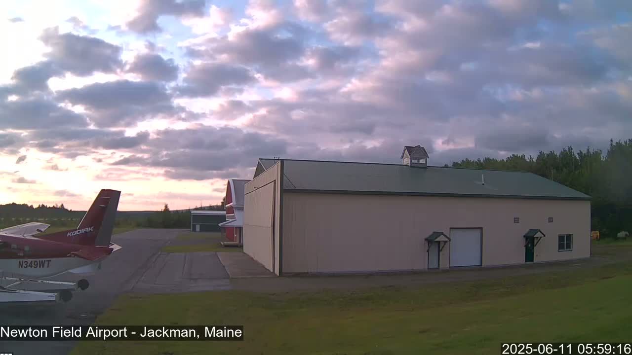 A small airplane sits on an airfield next to a large hangar under a partly cloudy, dusky sky.