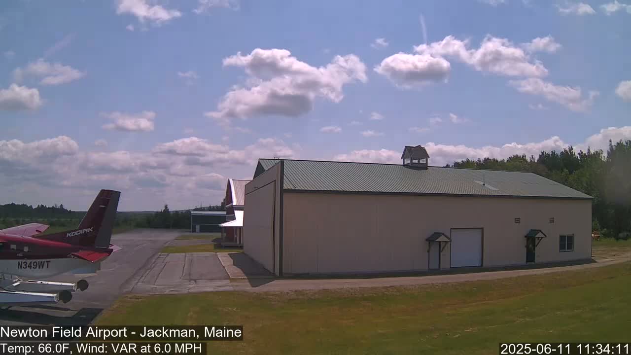 A red airplane is parked on an airfield next to a large beige hangar under a partly cloudy sky.