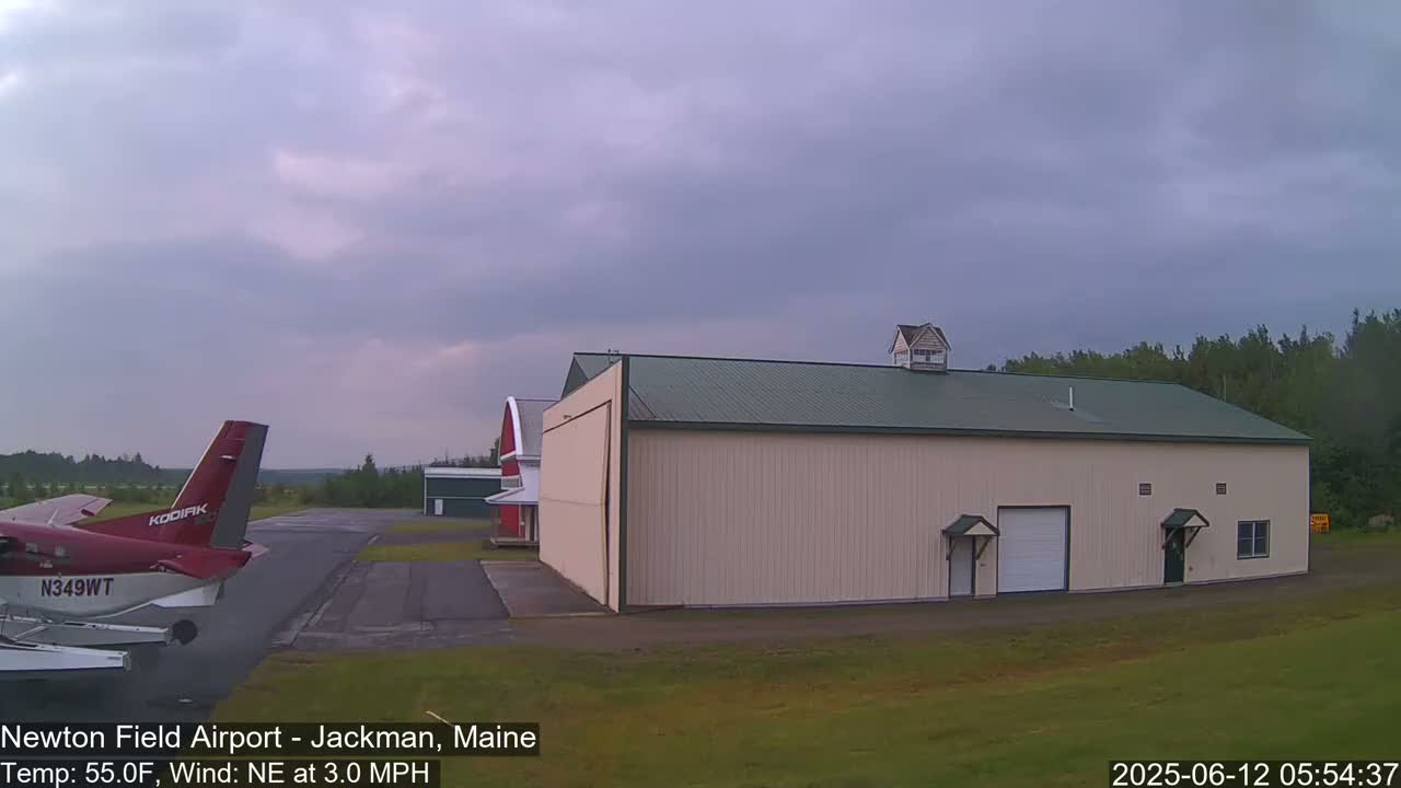 A red airplane is parked on an airfield next to a large beige hangar under an overcast sky.