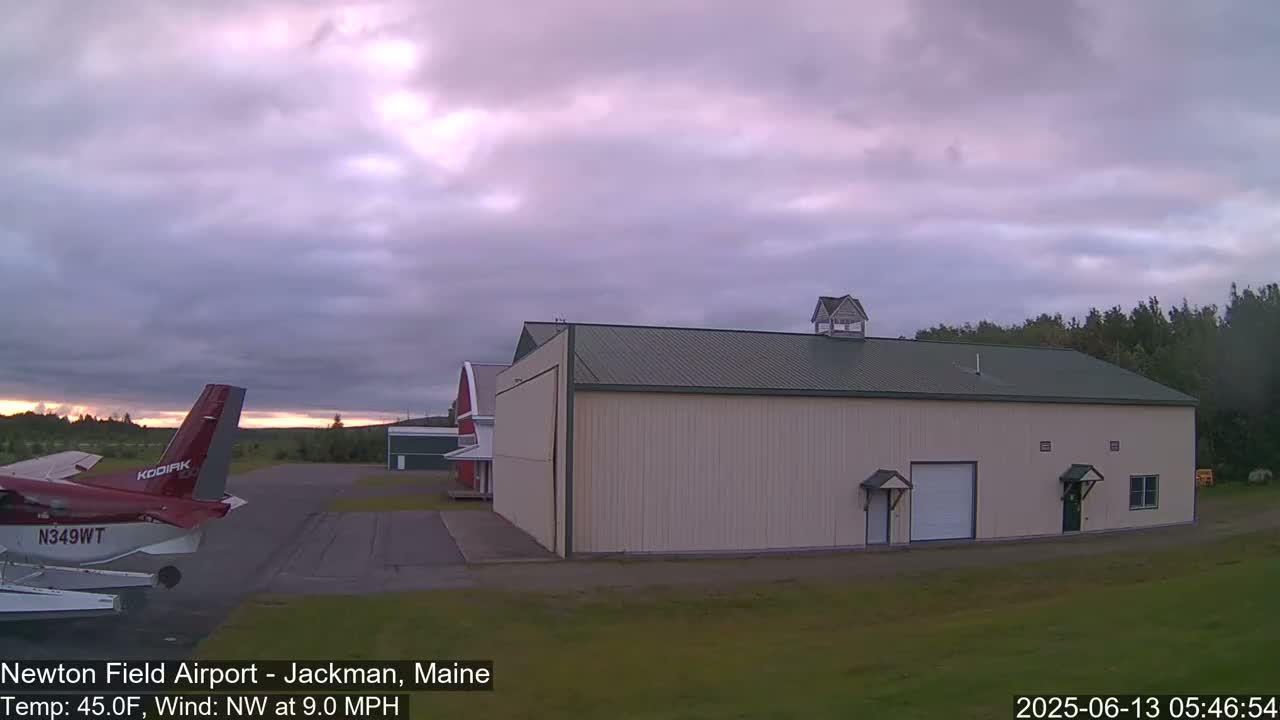 A red airplane sits on a tarmac near a large tan building under a mostly cloudy, dusky sky.