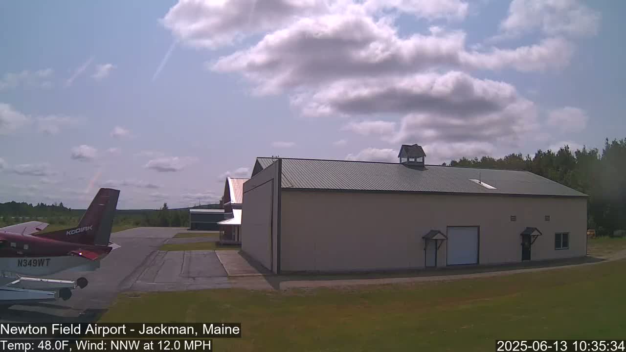 A small airplane is parked on an airfield next to a large hangar under a partly cloudy sky.