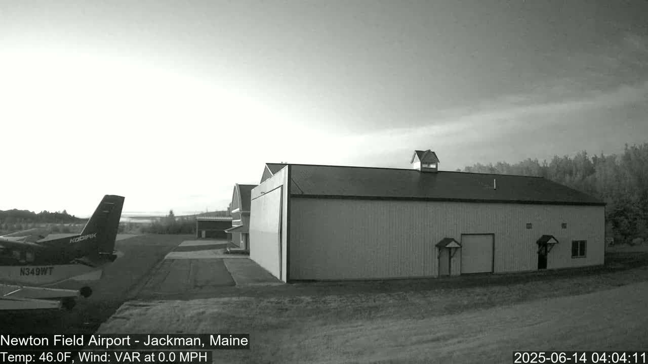 A small airplane is parked on an airfield near a large hangar under a clear, light sky.