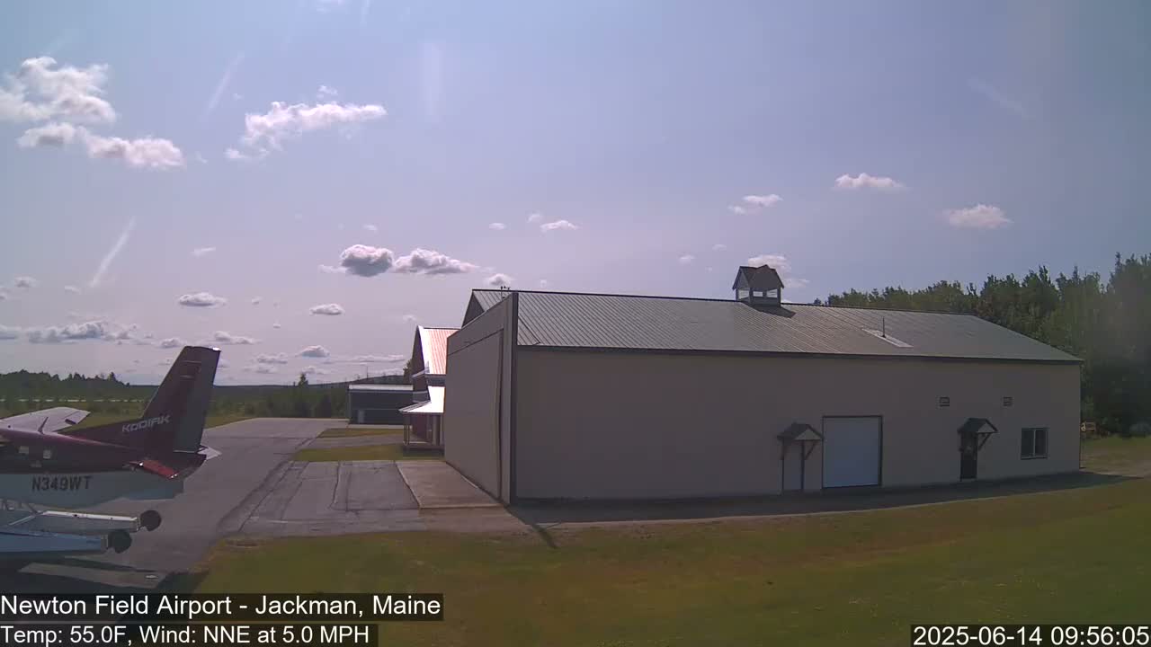 A small airplane is parked on an airfield near a large hangar under a partly cloudy sky.
