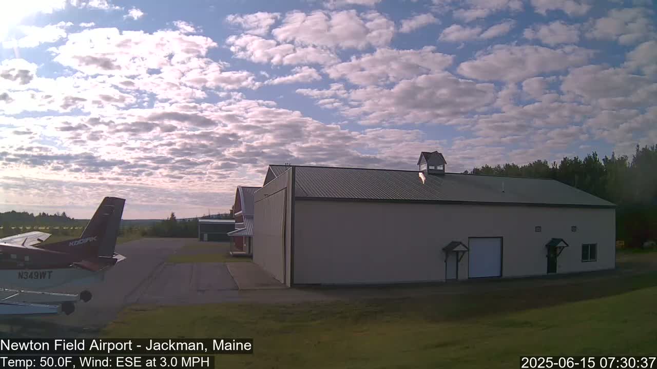 A small airplane sits on an airfield near a large tan hangar under a partly cloudy sky.