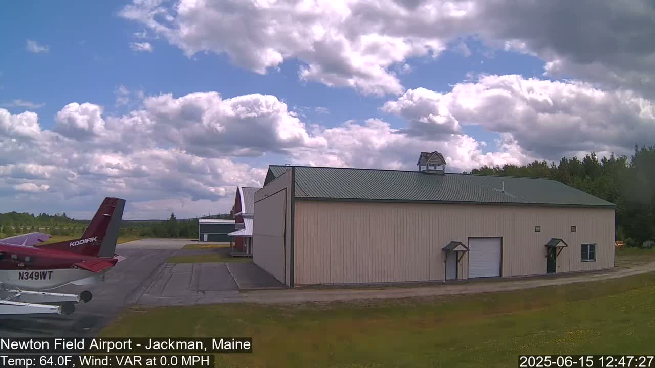 A red airplane is parked on an airfield near a large beige hangar under a partly cloudy sky.