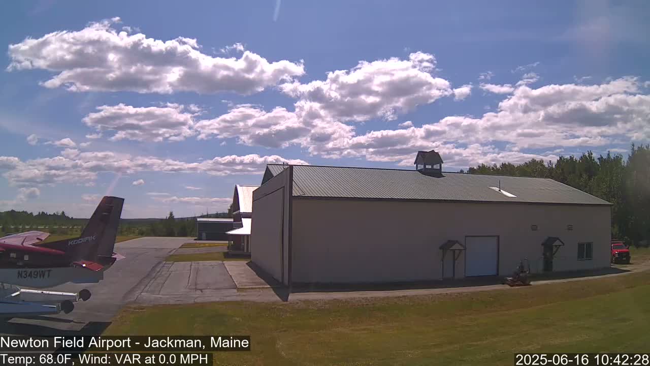 A small, red airplane is parked on an airfield next to a large, light-beige hangar under a partly cloudy sky.