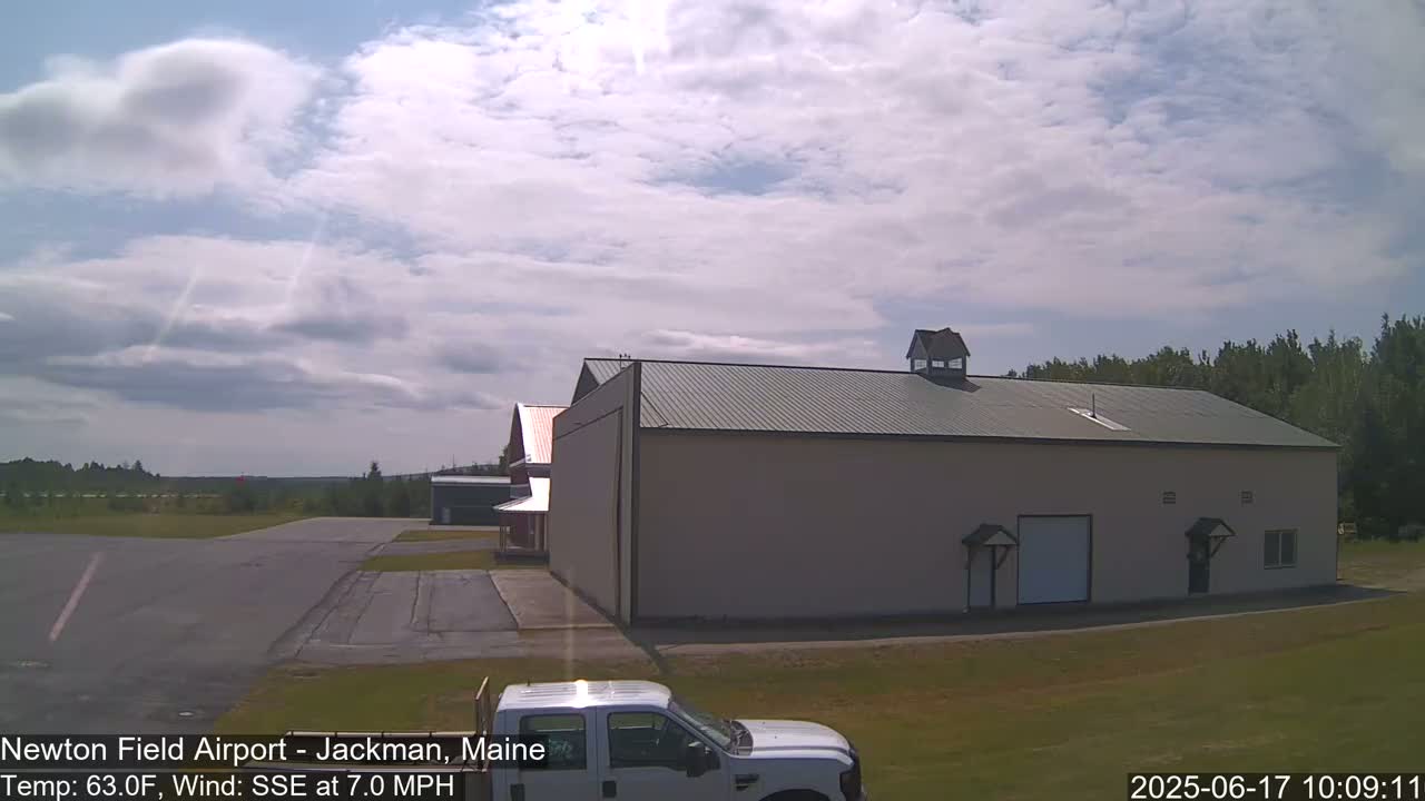 A partly cloudy sunny day at an airfield shows a large tan hangar, smaller red building, and a white pickup truck parked in the foreground.