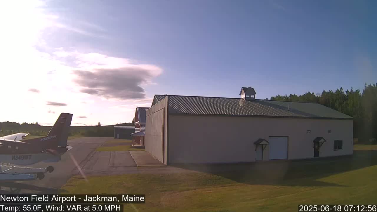 A small airplane is parked on an airfield next to a large hangar under a mostly clear sky with a few scattered clouds.