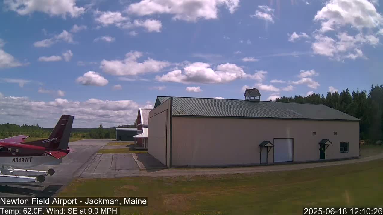 A red airplane is parked on an airfield next to a large beige hangar under a partly cloudy sky.