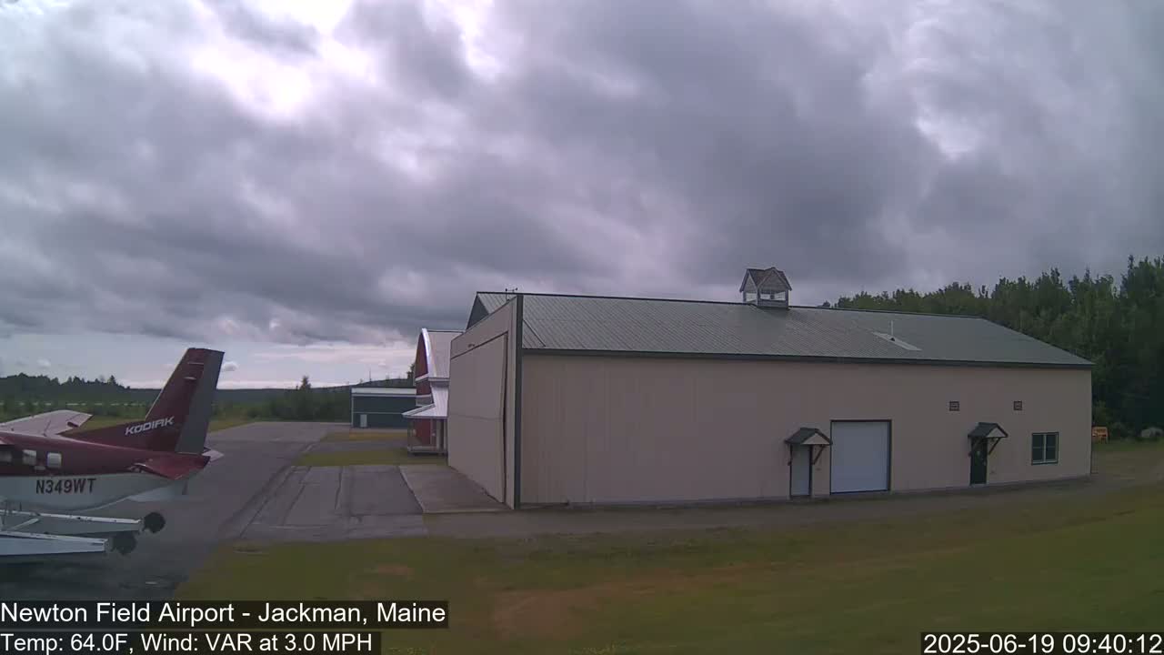 A red airplane is parked on an airport tarmac next to a large tan hangar under a cloudy sky.