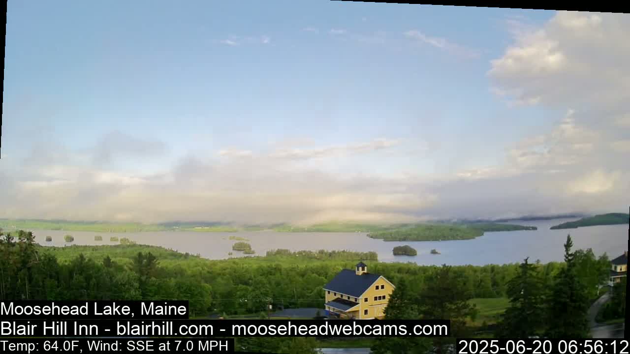 A yellow building sits atop a verdant hill overlooking a calm lake dotted with islands under a partly cloudy sky.