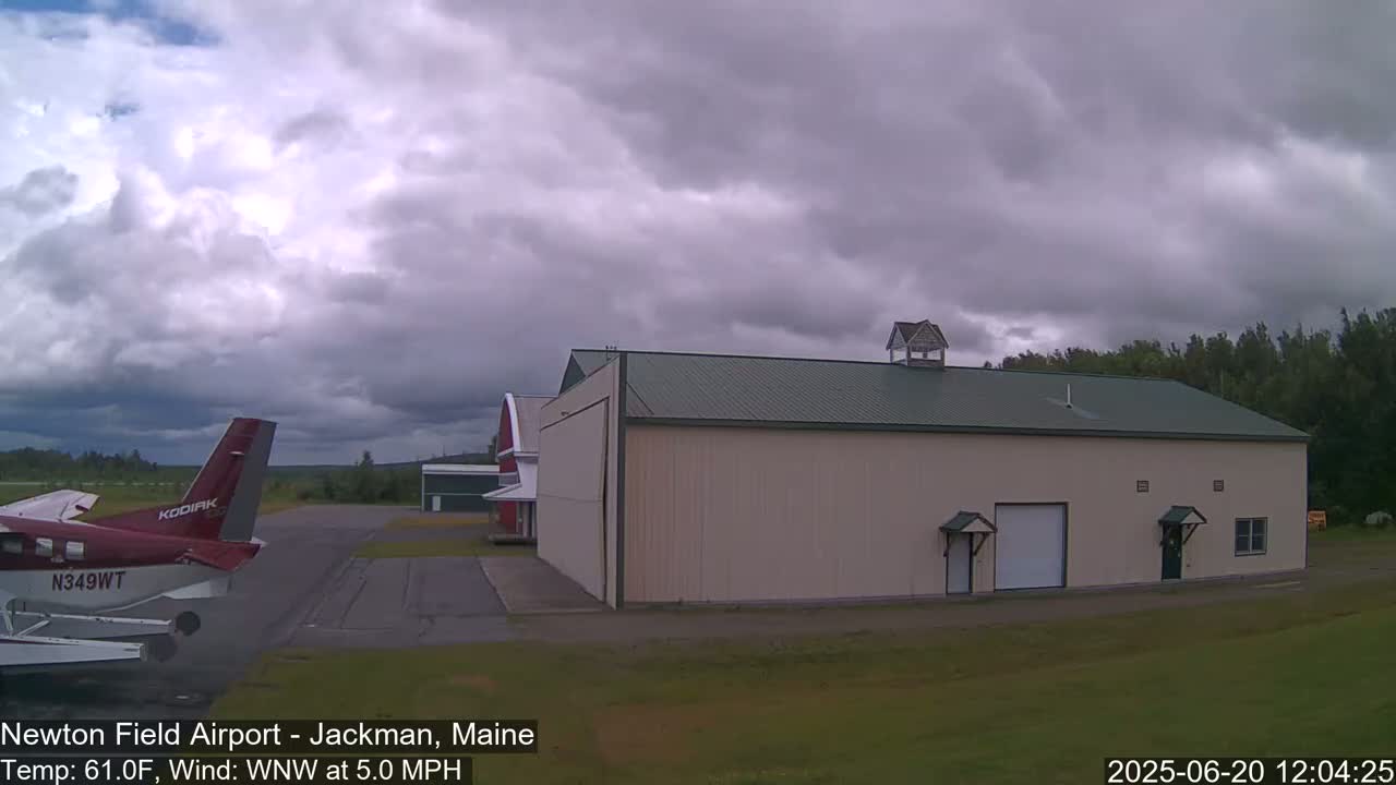 A red and white airplane is parked on an airfield next to a large tan hangar under a cloudy sky.