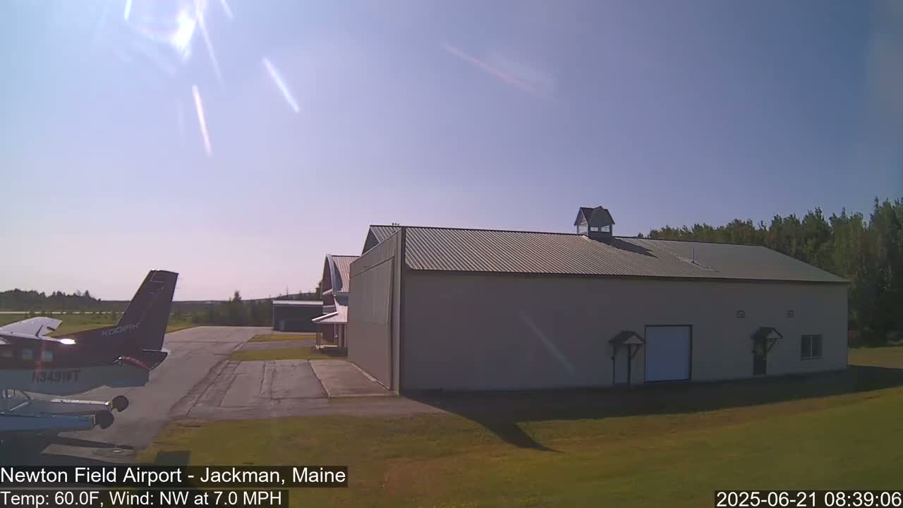A small airplane sits on a tarmac next to a large hangar under a sunny sky.