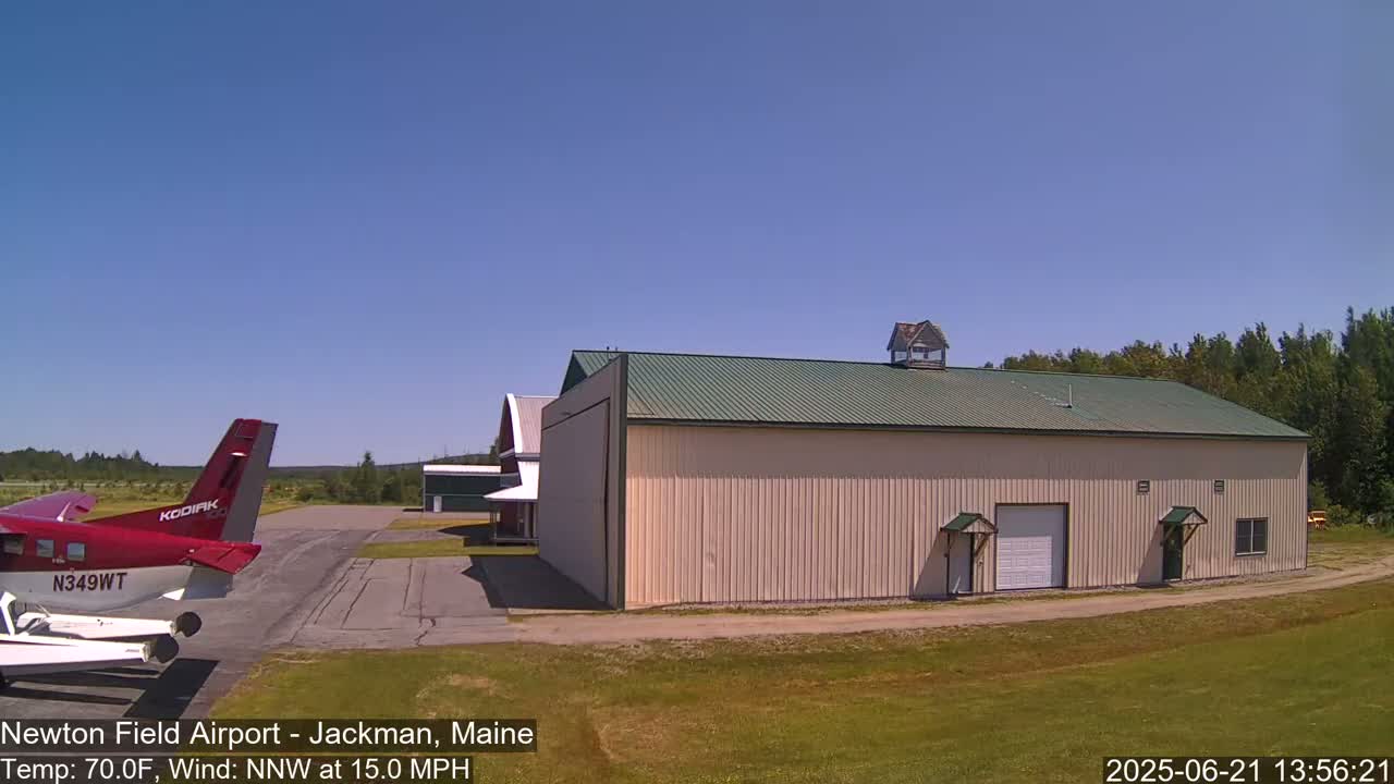 A red airplane is parked on a tarmac next to a large beige hangar under a clear blue sky.
