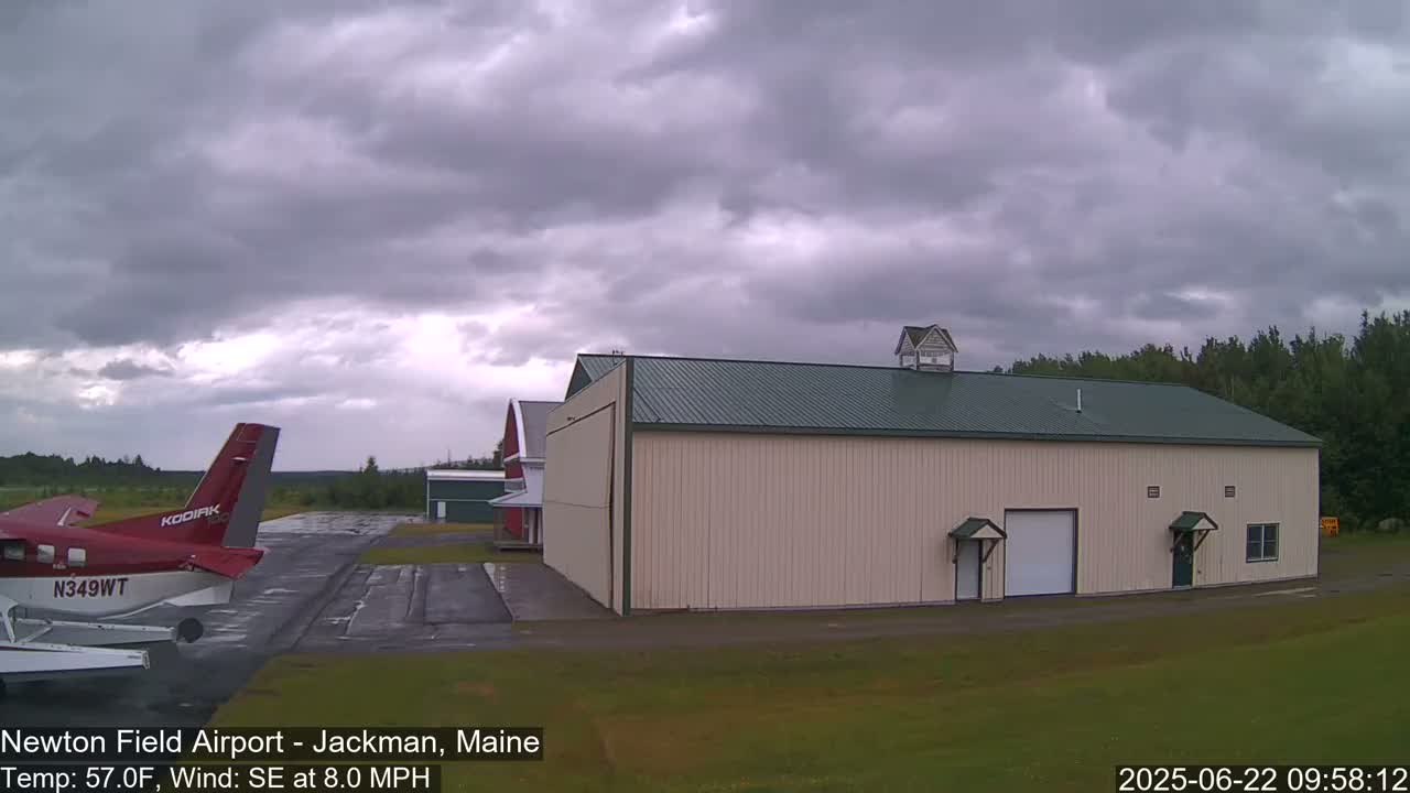 A red and white seaplane sits on a tarmac next to a large beige hangar under an overcast sky.