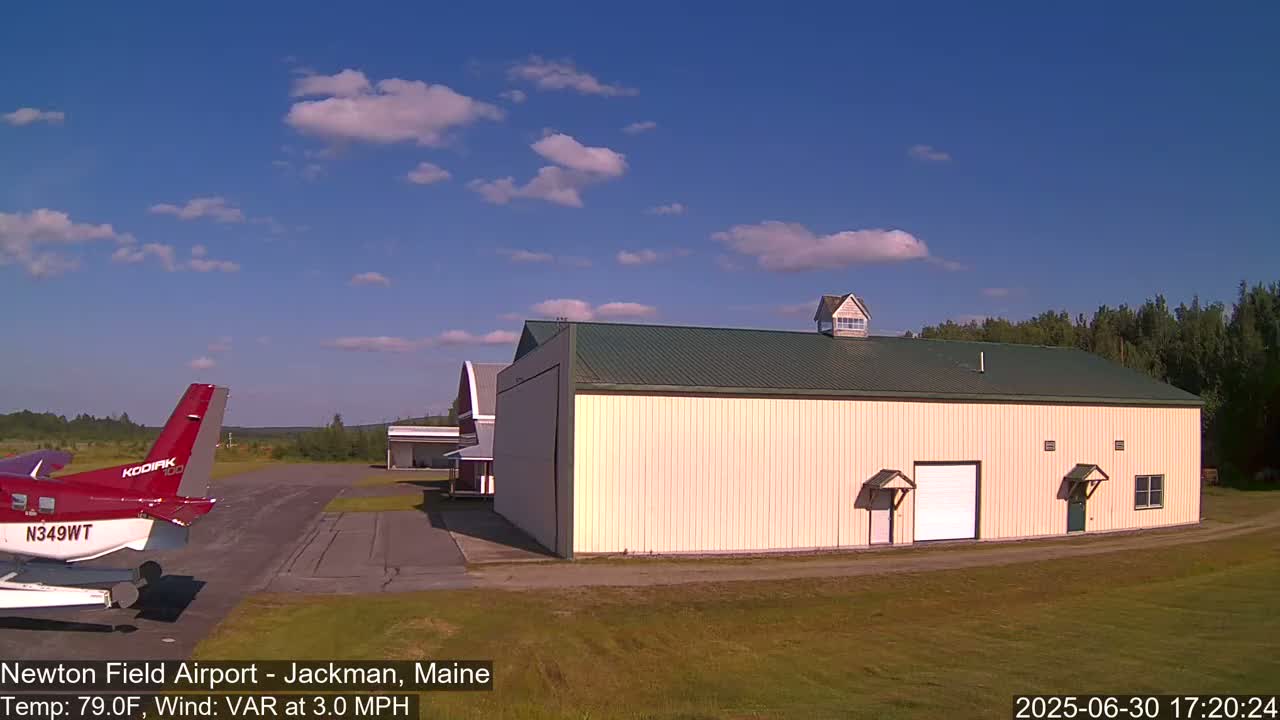 A red airplane sits on a grassy airfield next to a large tan hangar under a mostly sunny sky.