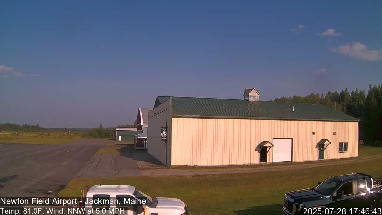 A large tan building with a green roof sits on a grassy area next to a paved runway, under a mostly clear blue sky.