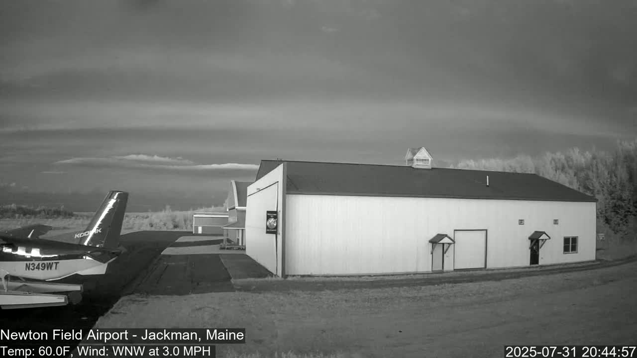 A small airplane is parked on an airfield next to a large white hangar under a mostly cloudy sky with a light wind.