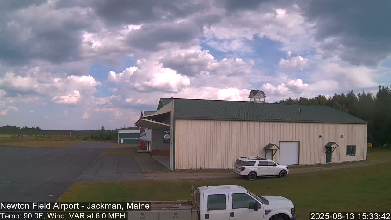 A large beige hangar with a green roof sits on an airfield under a partly cloudy sky, with a few vehicles parked nearby.