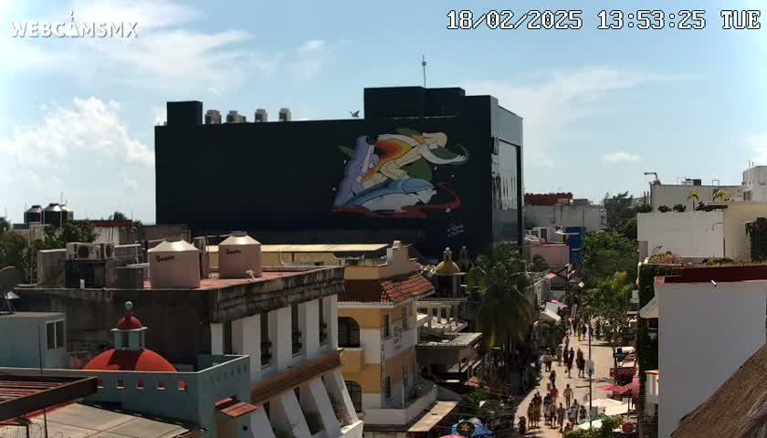 A city street scene on a sunny day, featuring a large building with a mural of a person surfing, and numerous other buildings surrounding it.