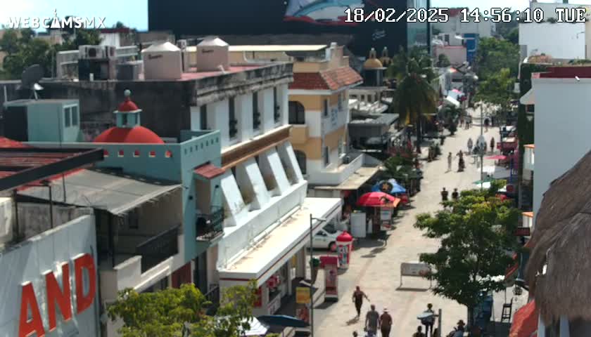 A sunny day reveals a pedestrian street lined with colorful buildings, some with shops and awnings, with people walking and cars parked along the sides.