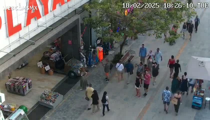A pedestrian walkway outside a brightly lit store is filled with many people, and a vintage car is parked nearby under a partly cloudy sky.