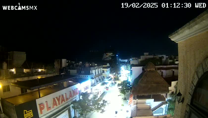 A nighttime, high-angle view of a city street, illuminated by streetlights, with buildings on either side and a thatched roof structure visible.