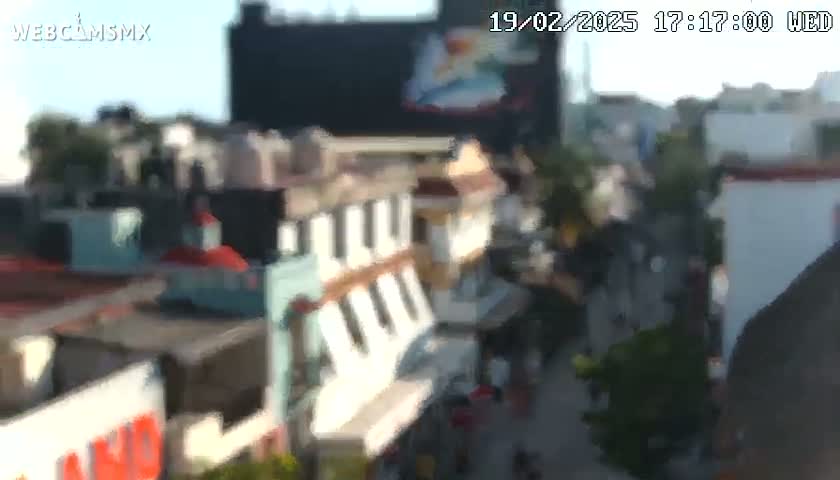 A blurry, high-angle view of a city street scene on a sunny day shows a collection of buildings with pedestrians walking along a street below.