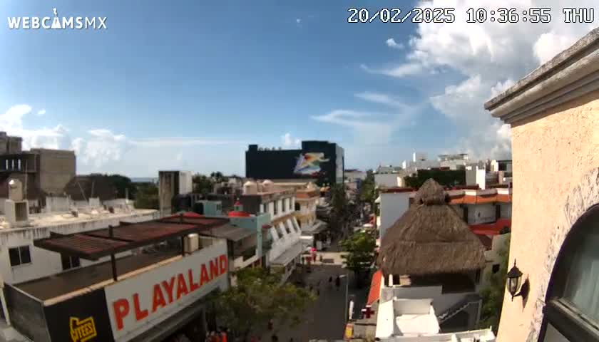 A mostly sunny, daytime view of a city street, showing a mix of buildings, including low-rise structures and taller buildings, with a thatched-roof structure visible in the foreground.