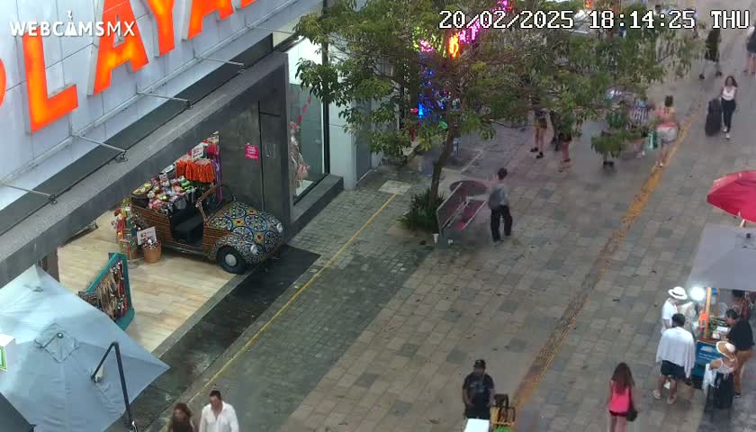 A high-angle view shows a paved pedestrian area with people walking past shops and a vendor's stall, under an overcast sky.
