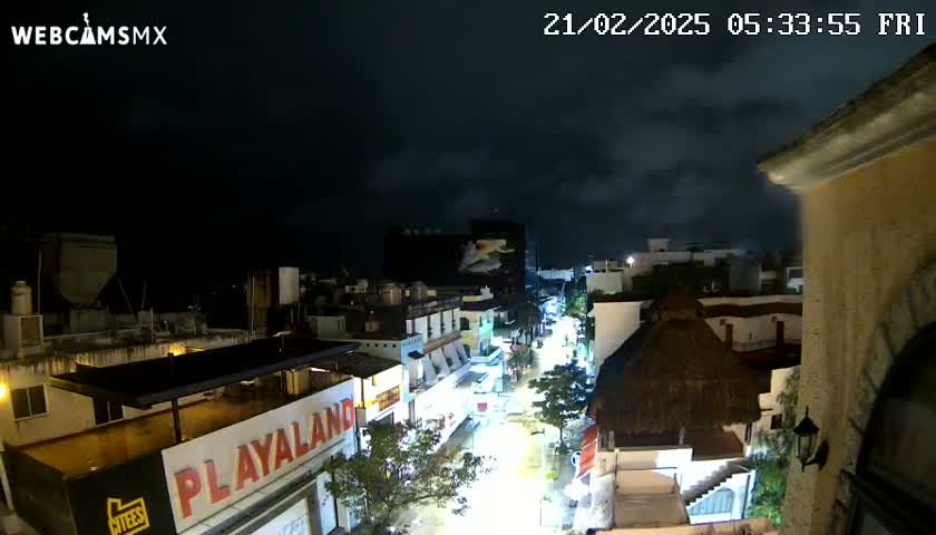 A nighttime view of a city street with buildings and palm trees under a cloudy sky.