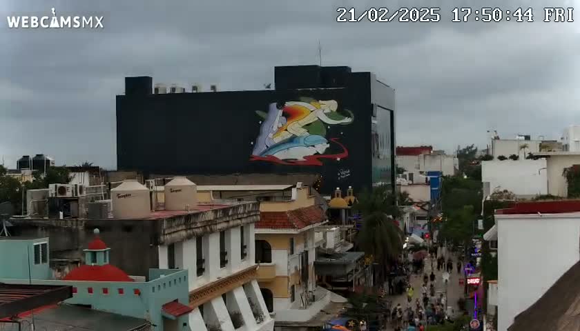 A city street scene under an overcast sky features a large building with a mural depicting stylized figures and aquatic life, surrounded by other buildings and pedestrians.