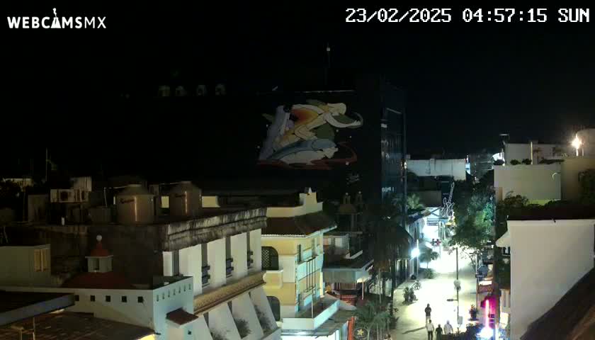 A nighttime view of a city street scene with buildings illuminated, a large mural on a building, and sparse pedestrian traffic.