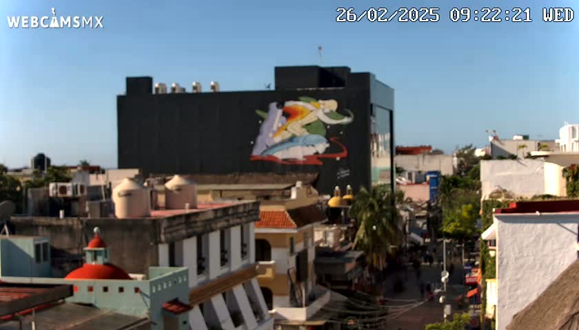 A city street scene on a sunny day, featuring a large building with a colorful mural, surrounded by smaller buildings with terracotta roofs.