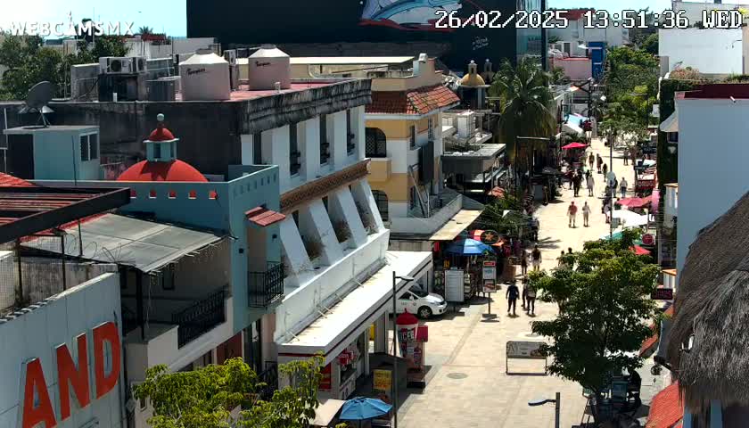 A sunny, pedestrian street scene shows multi-story buildings with various shops and restaurants, with people walking along the street.