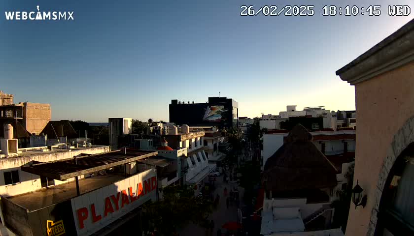 An aerial view of a city at sunset, showing buildings, rooftops, and a few people walking in the street, under a clear sky.