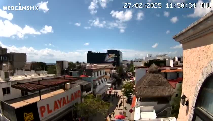 A partly sunny daytime view of a city street scene with buildings of varying heights, some with thatched roofs, and pedestrians walking below.