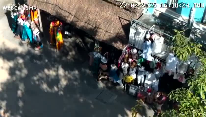 An aerial view shows a sunny outdoor market with people browsing and selling colorful clothing and goods under a thatched roof.