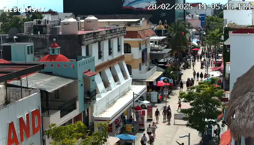 A sunny day reveals a bustling pedestrian street lined with colorful buildings, shops, and people walking amongst various street vendors.