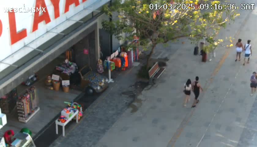 An overhead view of a sunny city street shows pedestrians walking past shops displaying various items, including clothing and souvenirs.