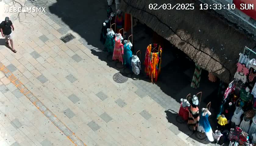 A man walks on a sunny paved sidewalk past a shop displaying colorful clothing on mannequins.