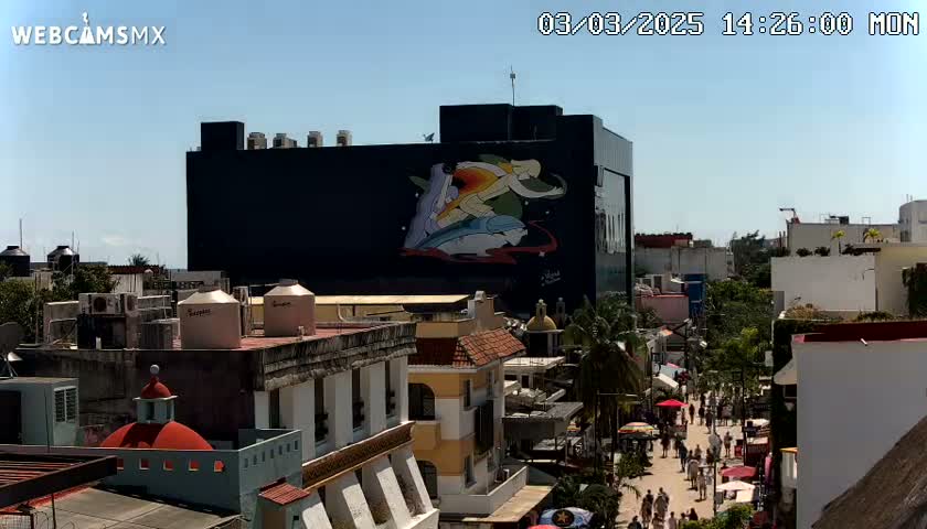 A city street scene on a sunny day, viewed from above, features a large building with a mural depicting figures and aquatic animals, surrounded by other buildings and pedestrians.