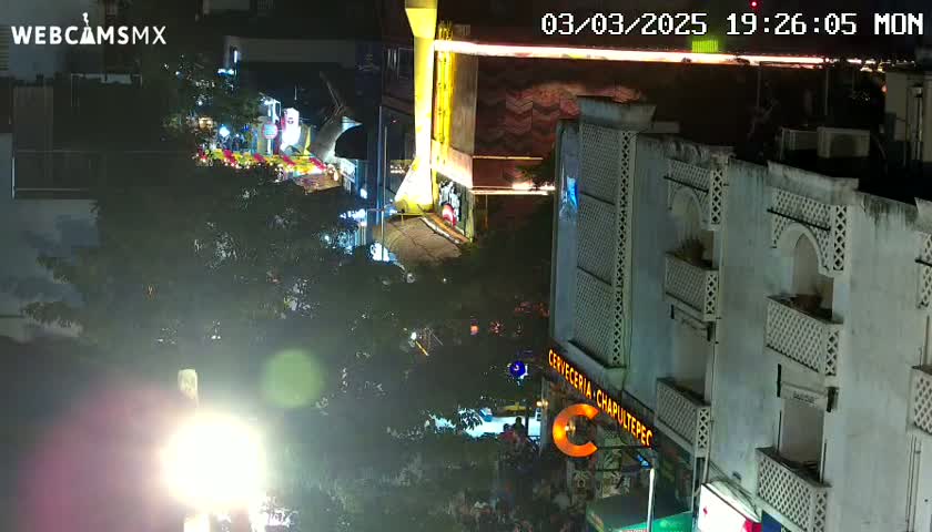 A nighttime aerial view of a city street with illuminated buildings and crowds of people gathered below, under a clear sky.