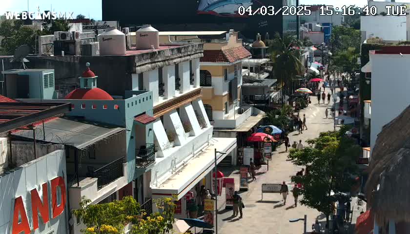 A sunny street scene shows pedestrians strolling along a pedestrian walkway lined with multi-story buildings, some with balconies, under a clear sky.