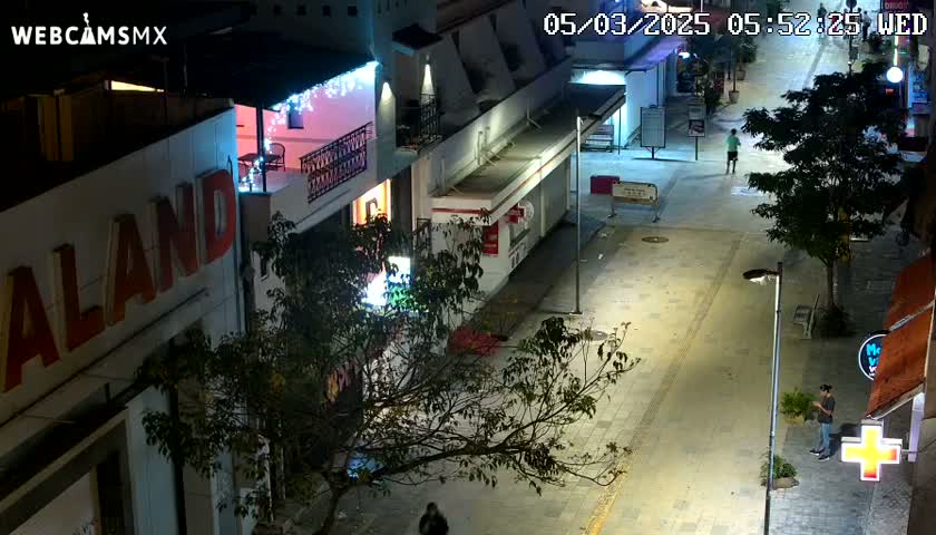 An aerial nighttime view of a mostly empty street with buildings and trees, under well-lit conditions.