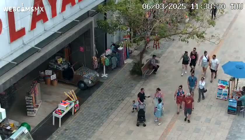 An outdoor paved walkway shows several people walking past shops and stalls, one of which has a decorated Volkswagen Beetle parked in front, under a partly sunny sky.