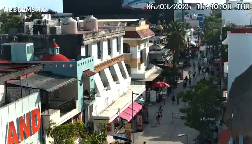 A sunny, high-angle view of a bustling pedestrian street lined with multi-story buildings, shops, and people walking.